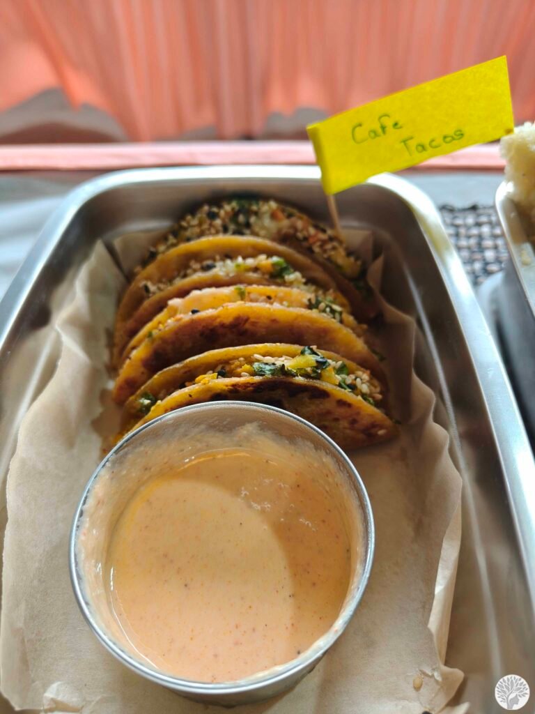 A close-up of Cafe Tacos lined up in a stainless steel tiffin lined with parchment paper, topped with a yellow label flag and served with a creamy dipping sauce.