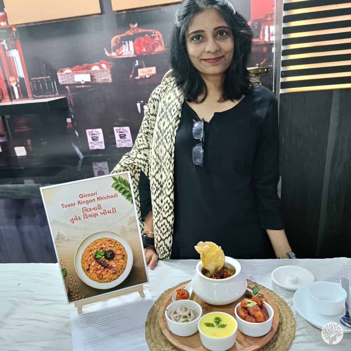 A smiling woman standing proudly behind a culinary competition display table featuring a traditional Kathiyawadi food platter and menu sign.