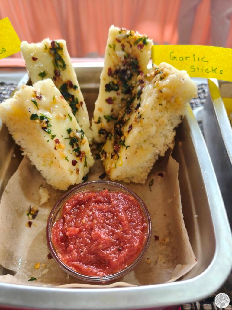 A close-up of four thick Garlic Cheese Sticks showing a spongy moong dal and rava dhokla texture, topped with melted cheese, herbs, and chili flakes, served with a red tomato dip.