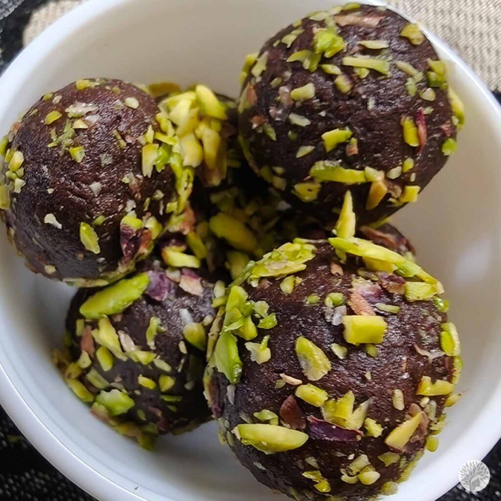 Four dark chocolate ragi cocoa truffles coated with chopped green pistachios, served in a small white bowl.