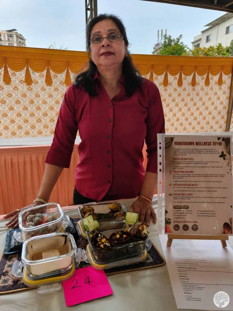 The author's mother standing proudly behind her cooking contest display table, featuring four clear tiffin boxes with her customized menu and a "Homegrown Wellness Tiffin" display placard at table number 24.