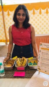 The author standing behind her beautifully arranged cooking competition table, displaying three stainless steel tiffin boxes with savory tacos, garlic bread dhokla, and dessert truffles next to a recipe placard.
