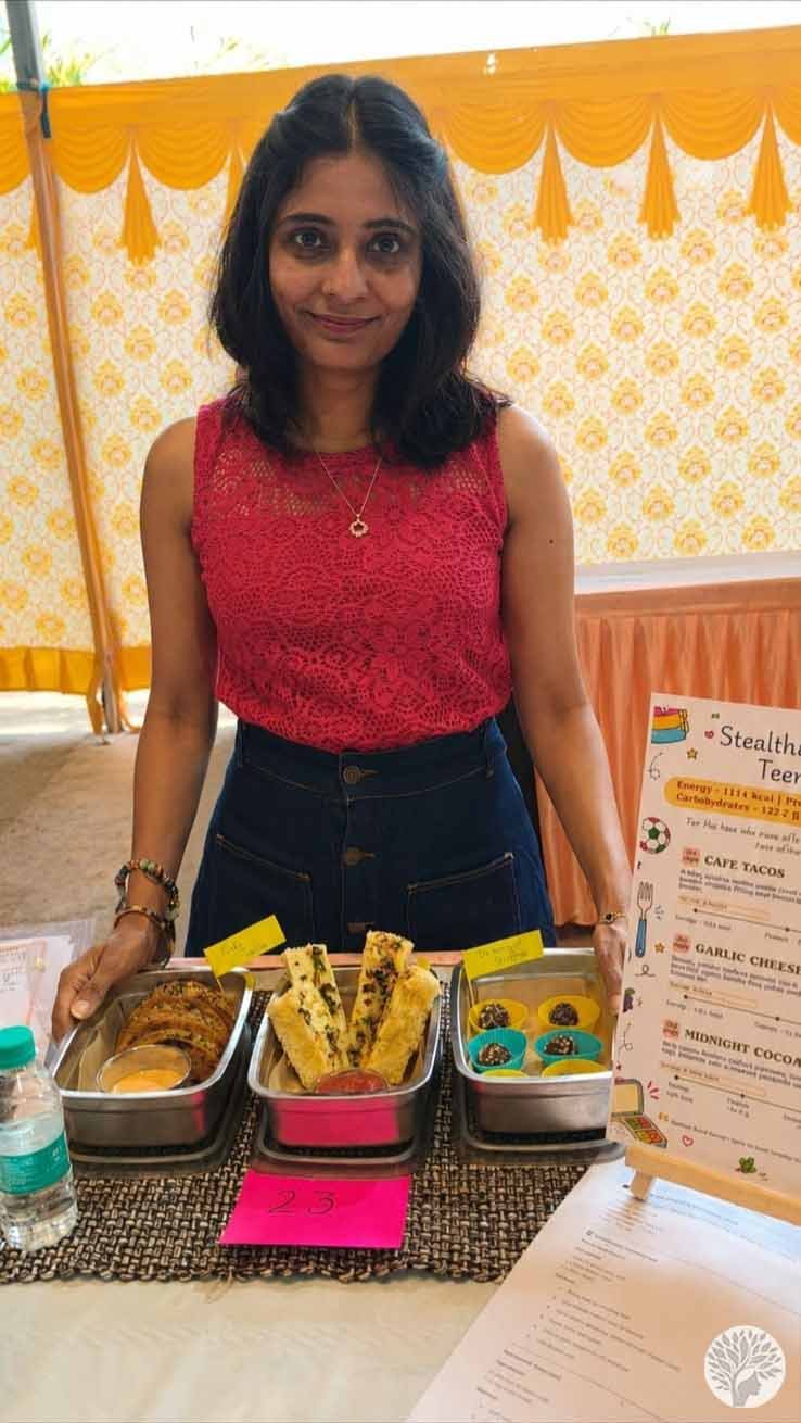 The author standing behind her beautifully arranged cooking competition table, displaying three stainless steel tiffin boxes with savory tacos, garlic bread dhokla, and dessert truffles next to a recipe placard.