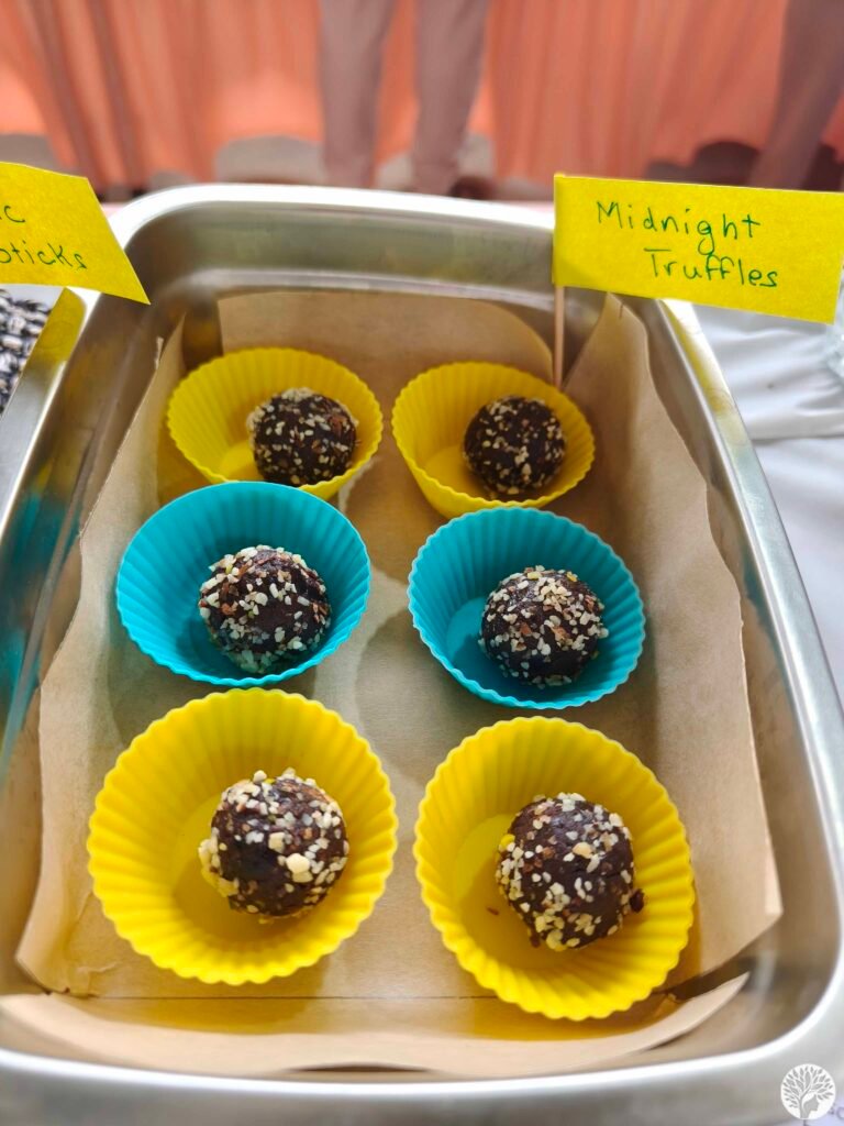 A close-up of six round Midnight Cocoa Truffles coated in crushed nuts, placed individually inside vibrant yellow and blue silicone liners within a tiffin container.
