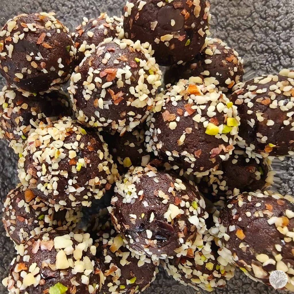 A glass bowl filled with a large batch of round dark chocolate ragi cocoa truffles heavily coated in finely chopped almonds and nuts.