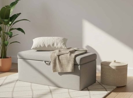 A minimalist light grey linen-textured storage ottoman bench with a lid, sitting on a patterned cream rug in a sunlit room. A knit throw blanket and a decorative pillow are placed neatly on top, and a woven lidded basket sits nearby, demonstrating hidden storage for visual silence.